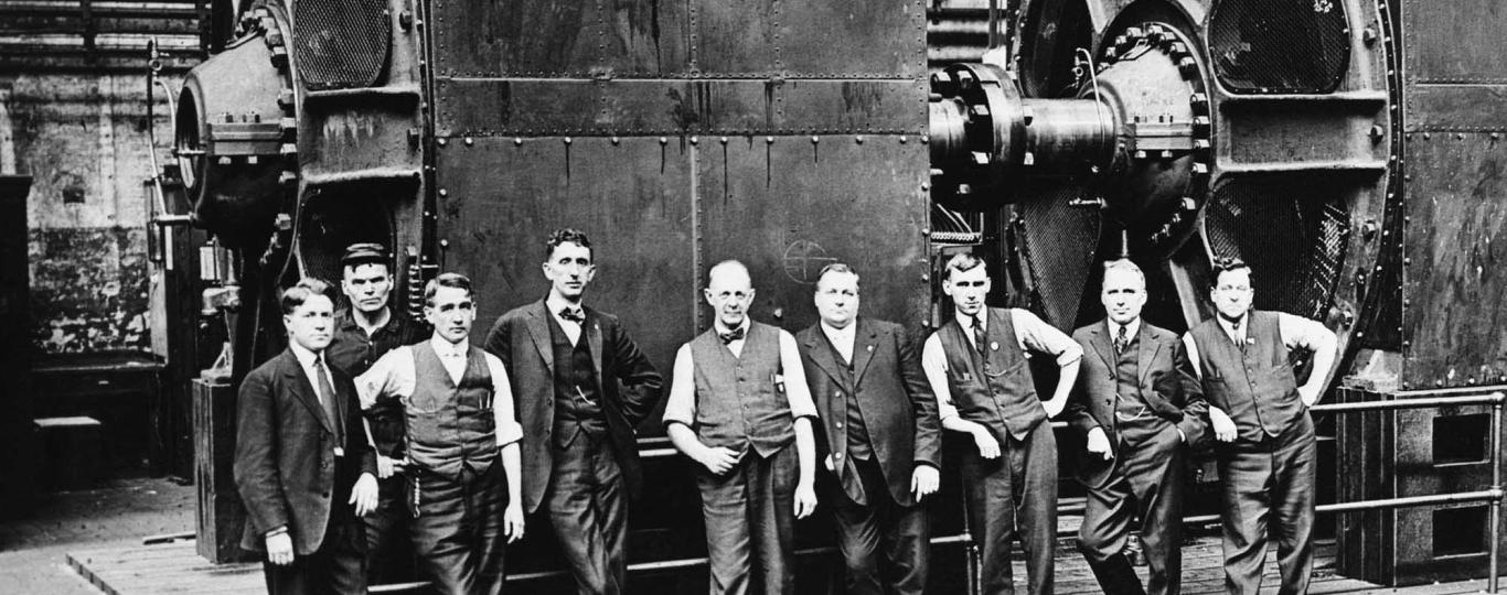 Mannen in pakken naast enorme dynamo's in een fabriek rond 1920