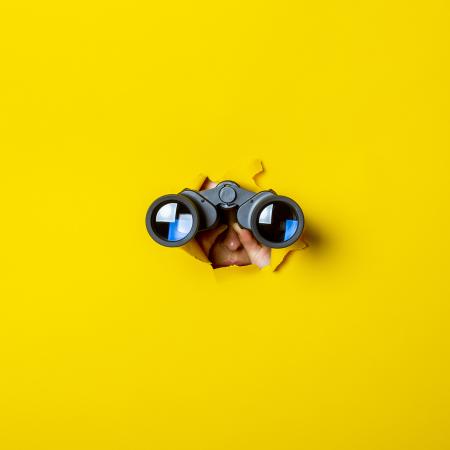 iemand met verrekijker gluurt door gat in gele wand