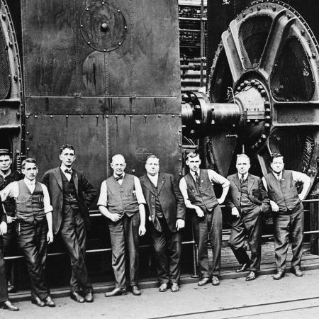 Mannen in pakken naast enorme dynamo's in een fabriek rond 1920